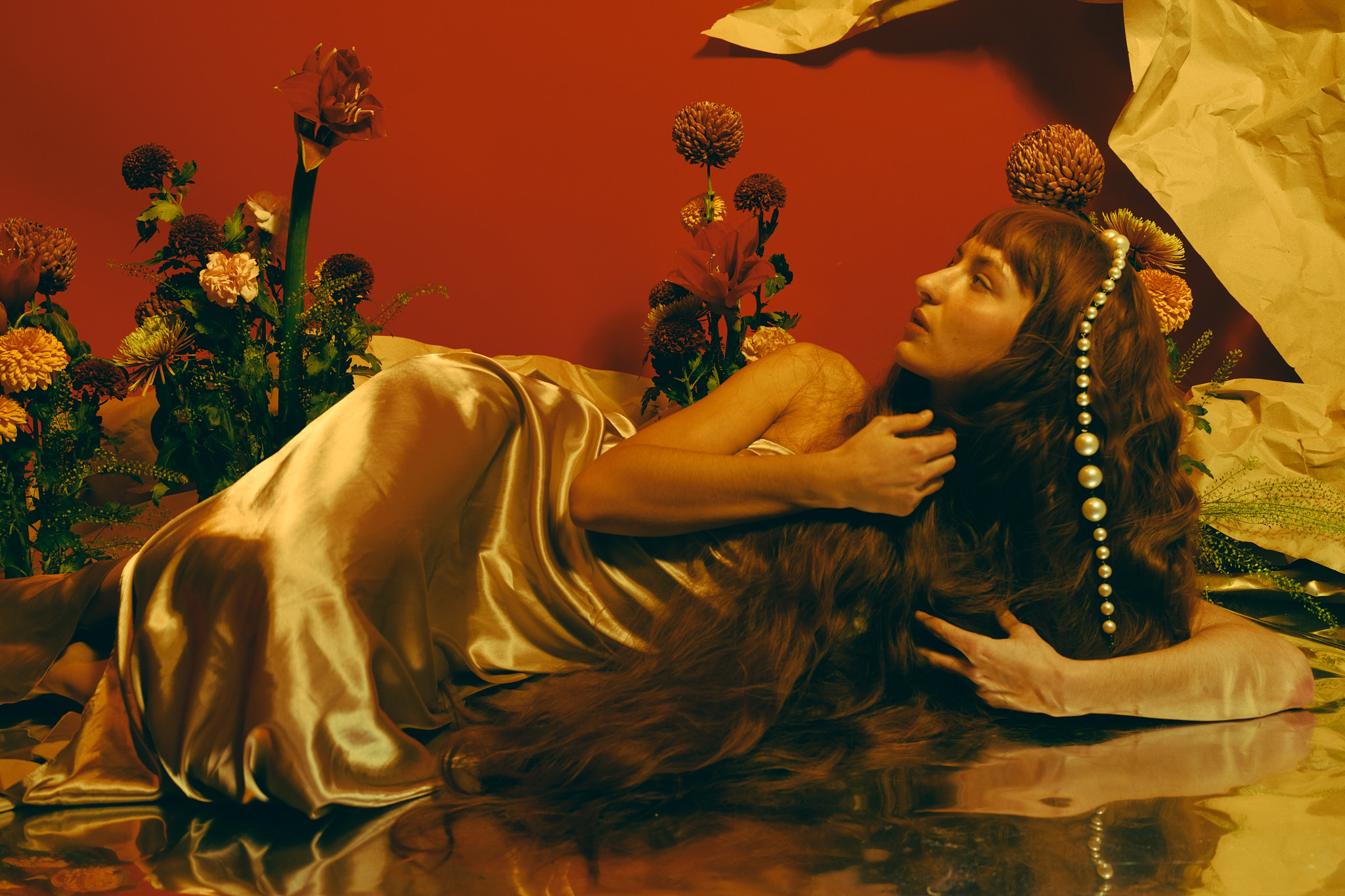 Model in gold satin reclining among flowers against a deep red backdrop